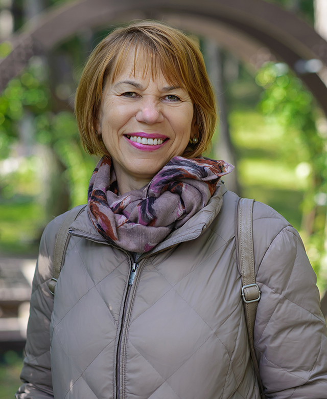 Natural Pharmaceuticals Vistan Vidutinio amžiaus moteris su trumpais rudais plaukais, šypsosi vilkėdama šviesią striukę ir spalvotą šaliką parke.