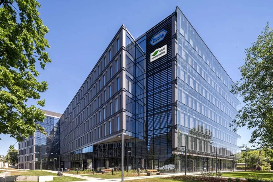 Natural Pharmaceuticals Vistan Modernus stiklinis biurų pastatas, apsuptas žalumos, su mėlynu dangumi fone.